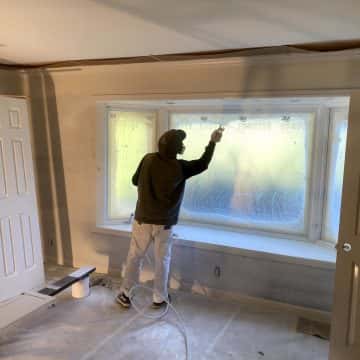 A man, surrounded by the faint scent of cigarette smoke, meticulously applies glossy oil paint to a window in a room.