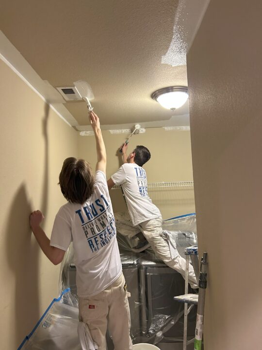 Two painters working on the ceiling of a room.