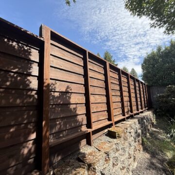 A tall wooden fence on a stone wall in Harvey's garden, greenery, and sky. Painted by Pearl Painters, Portland.