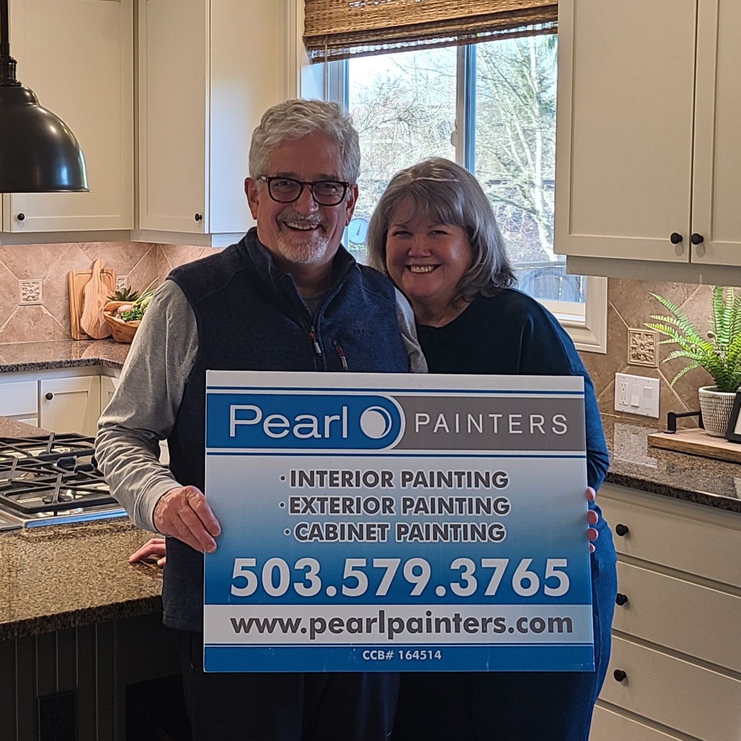 Two people in a kitchen hold a Pearl Painters sign with painting services and contact info, representing Portland painters.
