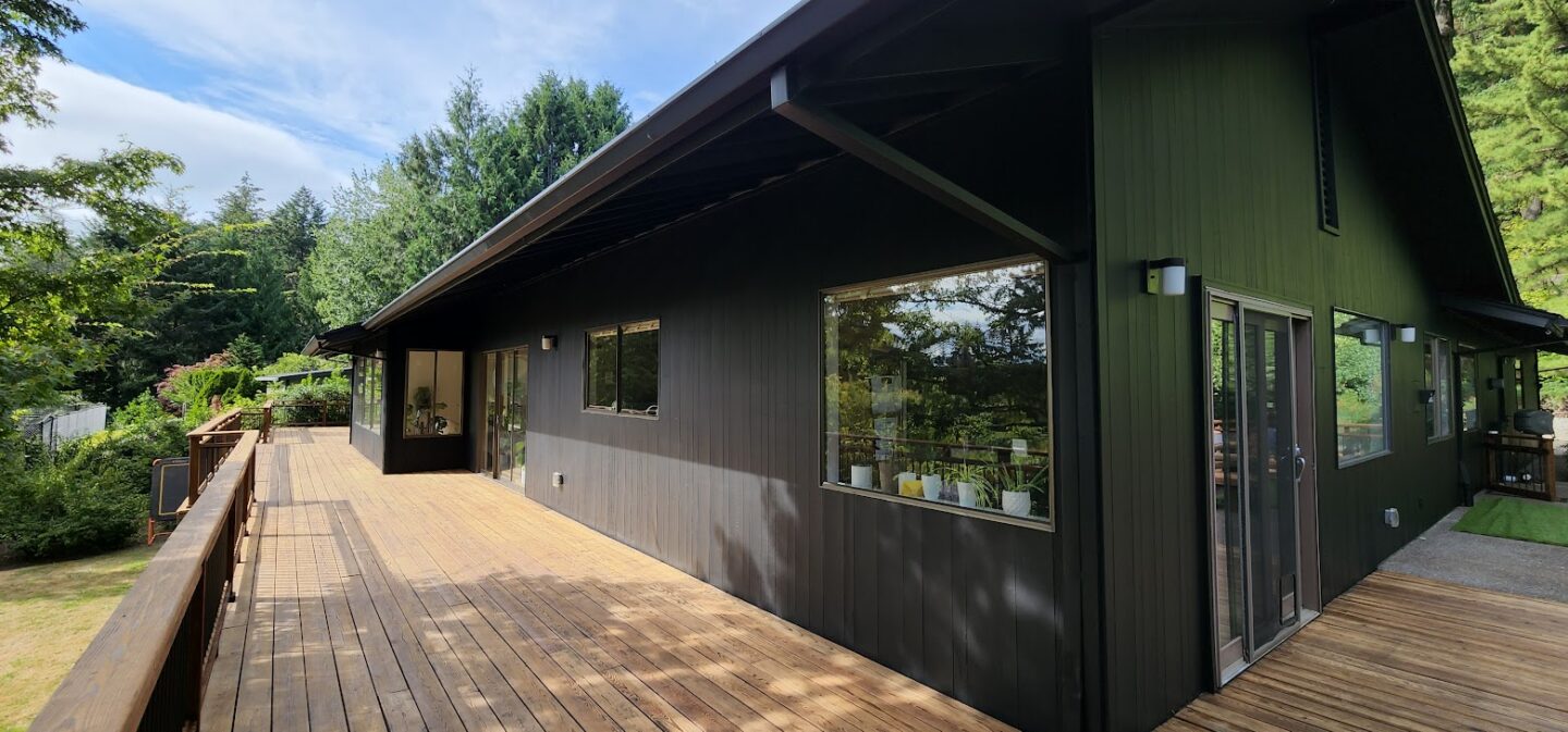 A modern dark-painted house in Portland with large windows and wood deck, painted by Pearl Painters, sits among trees.