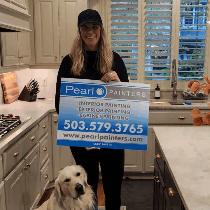 Woman in kitchen holds Pearl Painters painting sign with services and contact info; dog sits beside her. Likely in Portland.