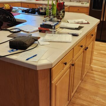 A cluttered kitchen island with books, papers, a purse, and various items spread out on top, set in a kitchen with wooden cabinets and flooring.