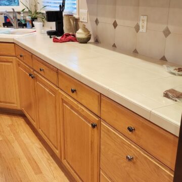 A clean kitchen with light wood cabinets, white countertops, and modern appliances. there are some red cloths and items on the counter.