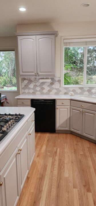 A modern kitchen featuring white cabinetry, a black dishwasher, a gas stove, and hardwood flooring with a view of greenery through a window.