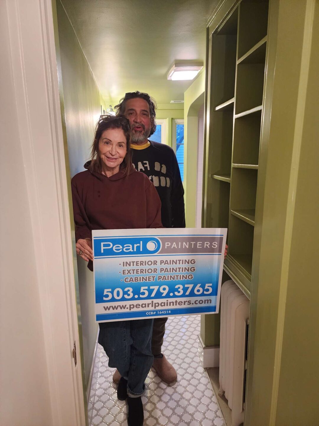 Two painters stand in a narrow hallway with green walls, holding a Pearl Painters sign advertising interior painting services in Portland. The sign displays their phone number and website.
