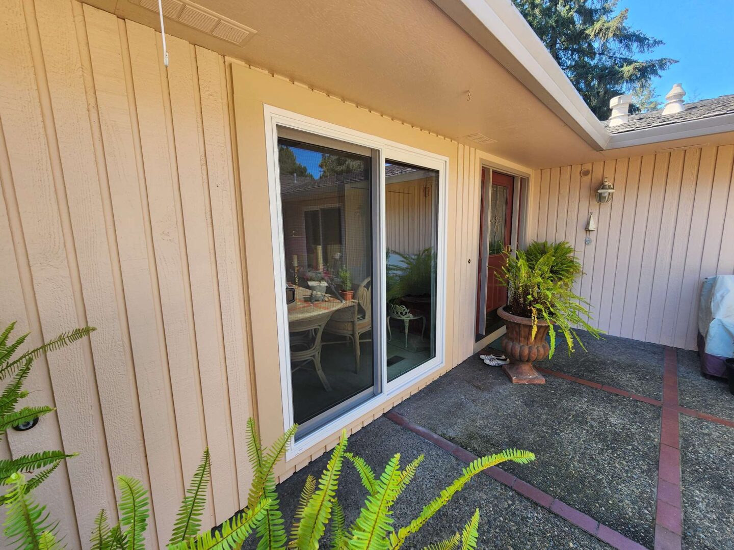 A beige exterior with vertical siding painted by Ferlitsch features a sliding glass door, red front door, potted ferns, and a patio area with indoor table and chairs—an April 2025 inspiration for Portland painters like Pearl Painters.