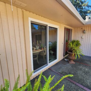 A beige exterior with vertical siding painted by Ferlitsch features a sliding glass door, red front door, potted ferns, and a patio area with indoor table and chairs—an April 2025 inspiration for Portland painters like Pearl Painters.