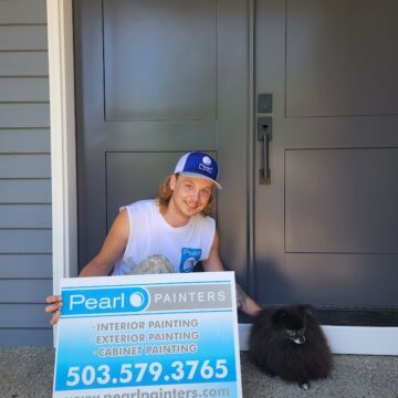 Person kneels by black dog at gray door, holding Pearl Painters Portland sign with painting services for Roberts neighborhood.