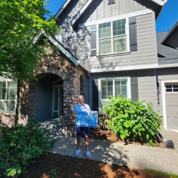 A painter stands by a modern two-story Portland home with a sign, lush plants visible—Pearl Painters July 2025 event.