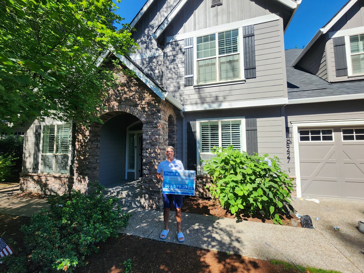 A painter stands by a modern two-story Portland home with a sign, lush plants visible—Pearl Painters July 2025 event.