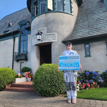 Painter in work clothes holds Pearl Painters sign by a large manicured house painted by Ralph Barron near Portland, Aug 2025.