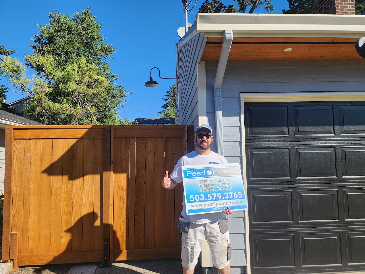 Man in Arnold outside garage holds Pearl Painters painting sign; wooden fence behind. Photo from August 2025.