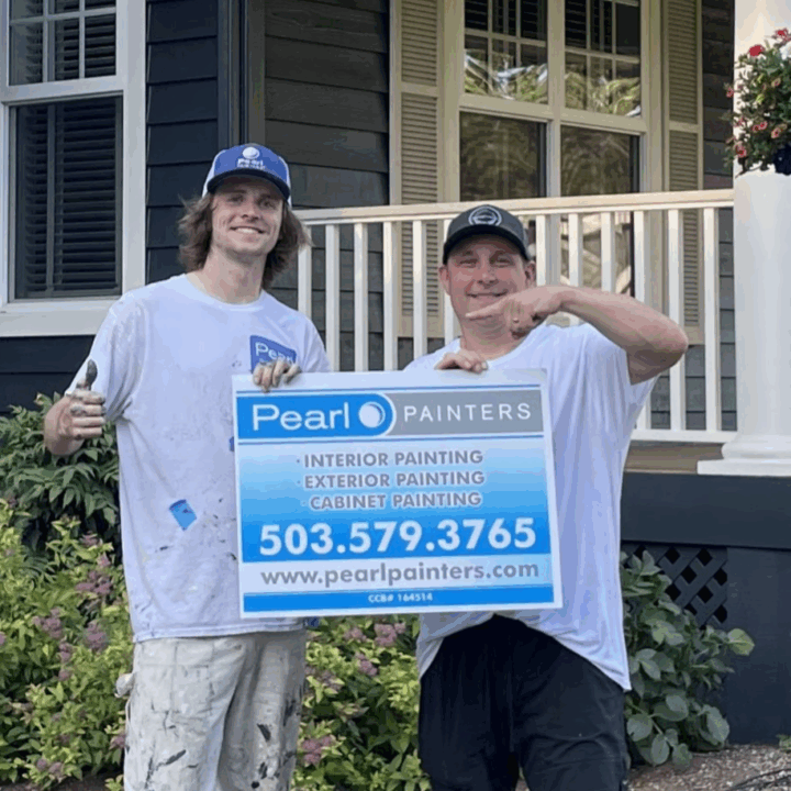 Two painters in work clothes hold a Pearl Painters sign for painting services and contact info in front of a Portland house.