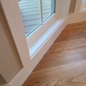 A professional painting of a corner of a window with interior trim and wooden floor.