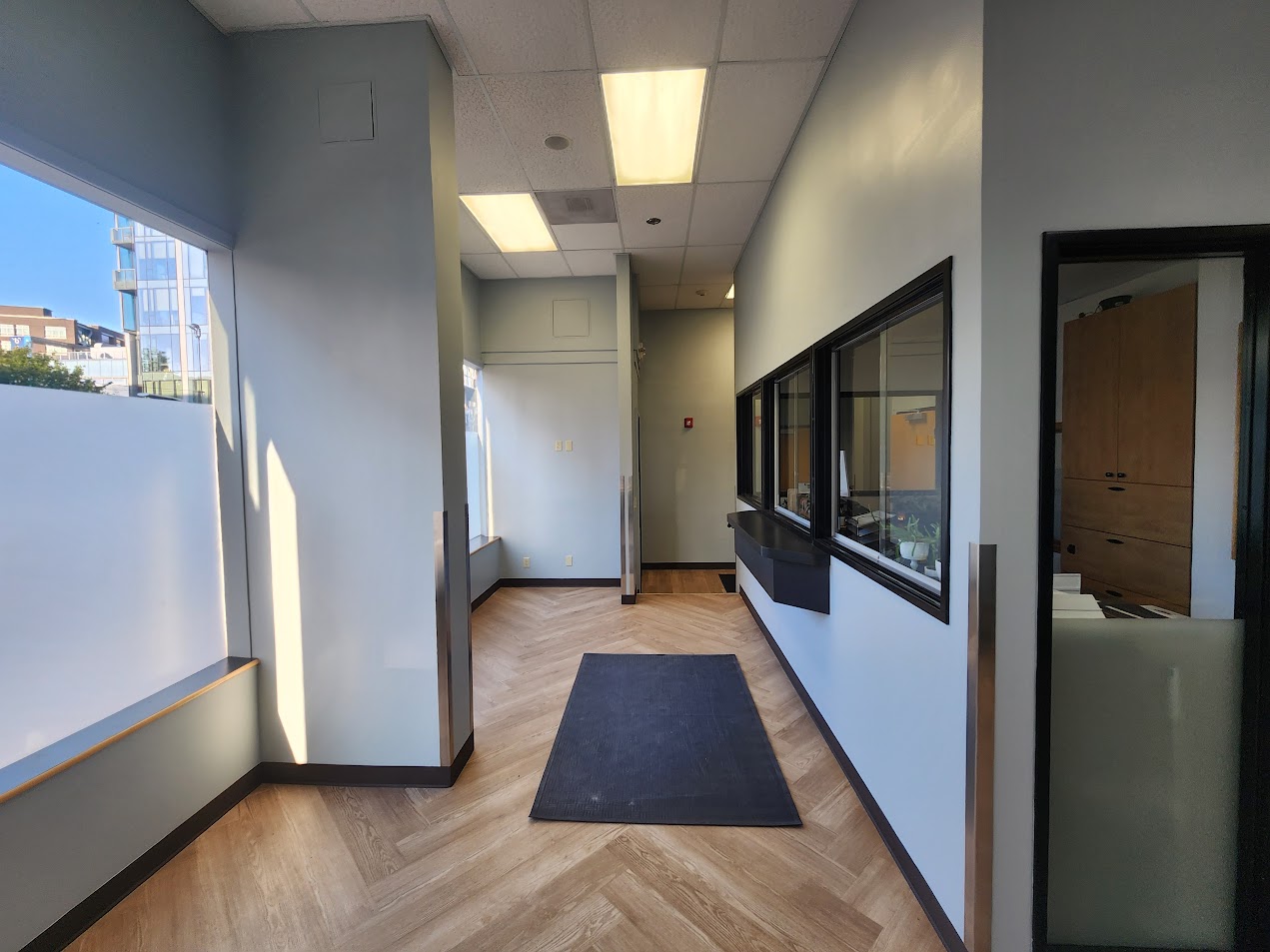 Modern office or clinic lobby in Portland with wood-patterned floor, black mat, large windows, and a reception window on the right. Natural light fills the space. Suitable for painting by Pearl Painters in June 2025.