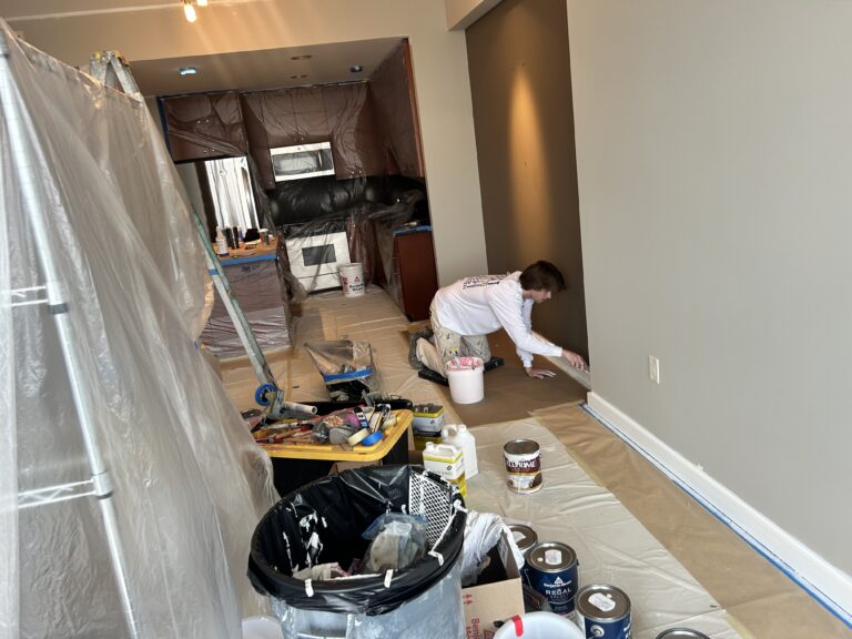 Person painting baseboards in a room under renovation, with plastic coverings, paint cans, and tools scattered around.