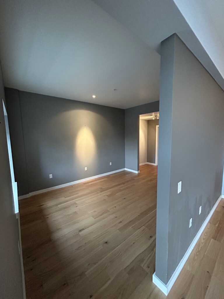 An empty room with gray walls, wood flooring, and a recessed lighting fixture. An entryway is visible in the background.