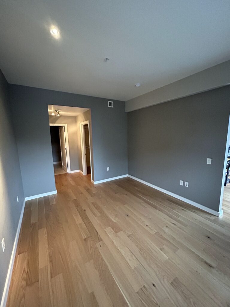 Empty room with gray walls and wooden floors, featuring a small ceiling light. A doorway leads to a hallway in the background.