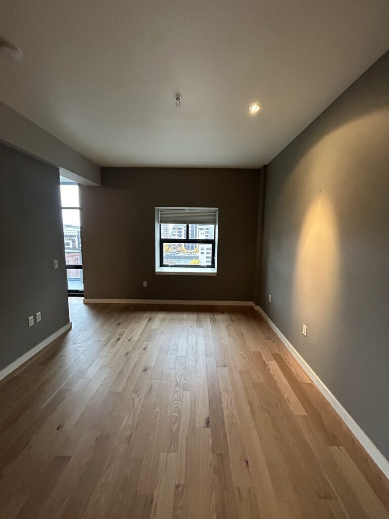 Empty room with wooden flooring, gray walls, and a small window showing a cityscape. Ceiling lights are on, illuminating the room.