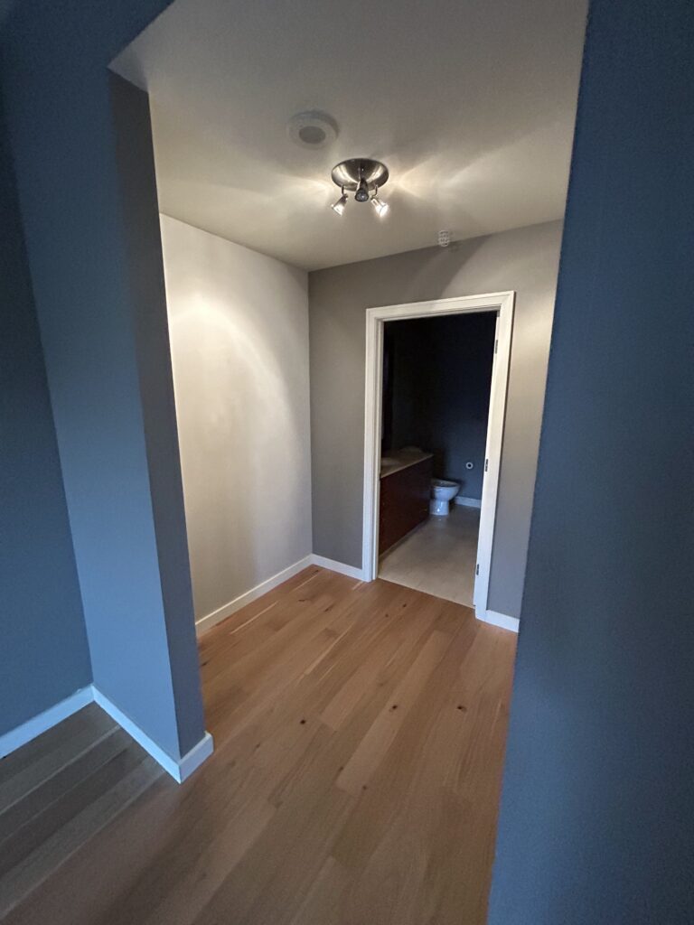 A small hallway with light wooden flooring leads to a bathroom. A light fixture is on the ceiling, and a toilet is visible through the bathroom door.