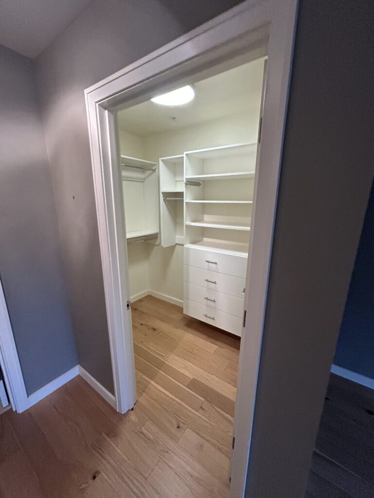 Open door view of a white walk-in closet with wooden flooring, featuring shelves and drawers.