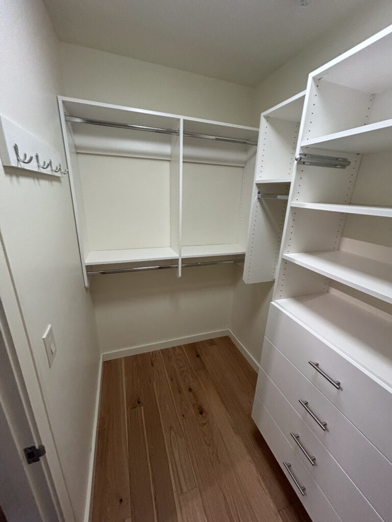 Empty walk-in closet with white shelves, drawers, and hanging rods. Light wood flooring and walls with a row of hooks on the left.