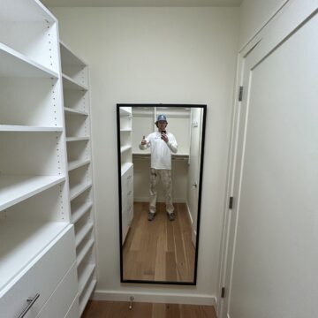 A person in a white outfit and hat takes a selfie in a full-length mirror inside a walk-in closet with white shelves and drawers.