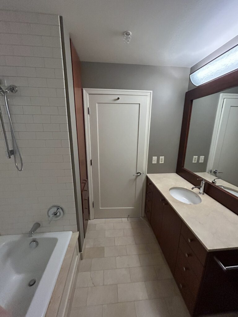 A modern bathroom with a bathtub, a white door, a large mirror above a sink, and wooden cabinetry. The walls are partially tiled, and the floor is light-colored.