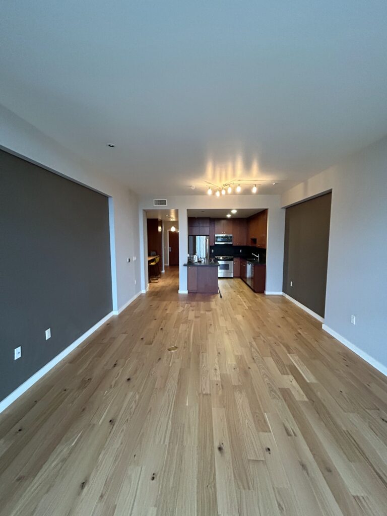 A modern, unfurnished apartment with wooden floors, recessed lighting, and an open kitchen. The room features large, dark-colored window shades.