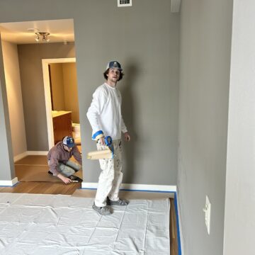 Two people work on a room renovation. One person stands with painting tools, while the other kneels on the floor. The room has grey walls and a protective sheet on the floor.