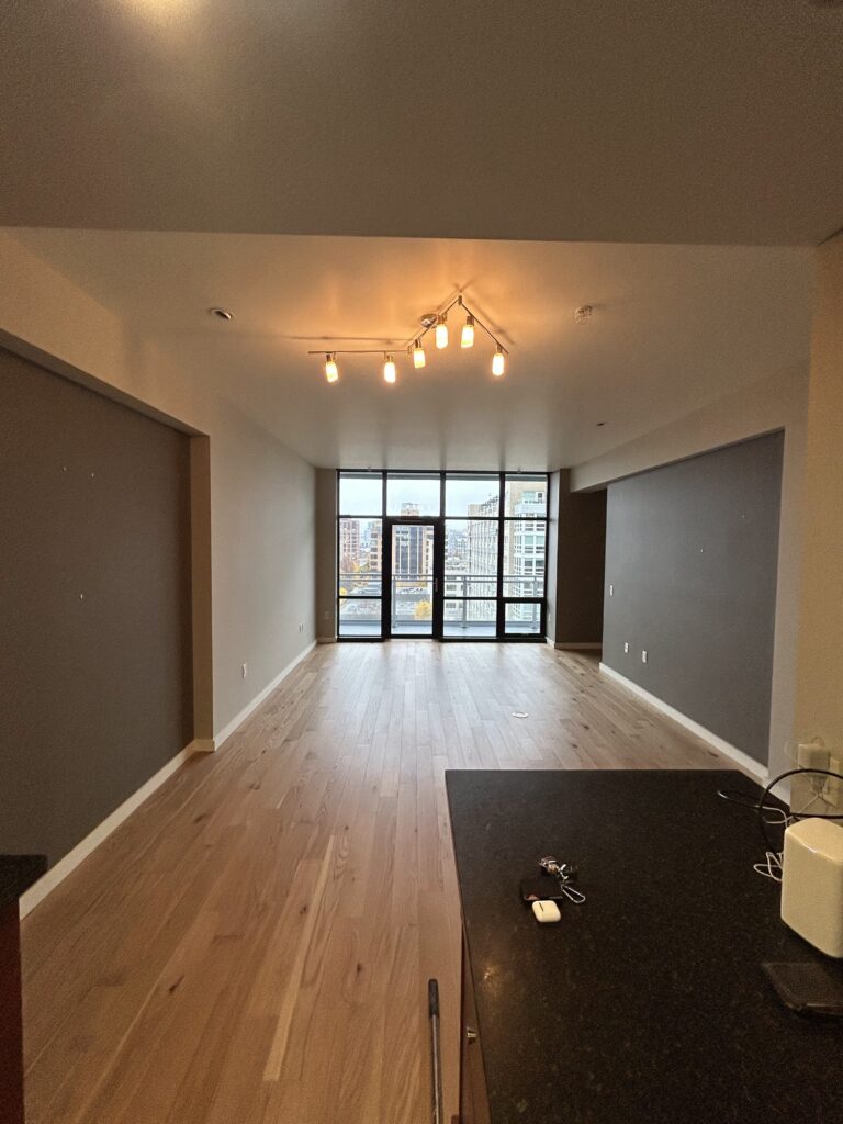 An empty room with wooden floors, dark accent walls, and a central light fixture. Large windows overlook a cityscape. A kitchen counter is partially visible in the foreground.