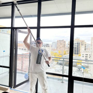 Person in white attire painting a ceiling with a roller on an extended pole in a room with large windows overlooking urban buildings.