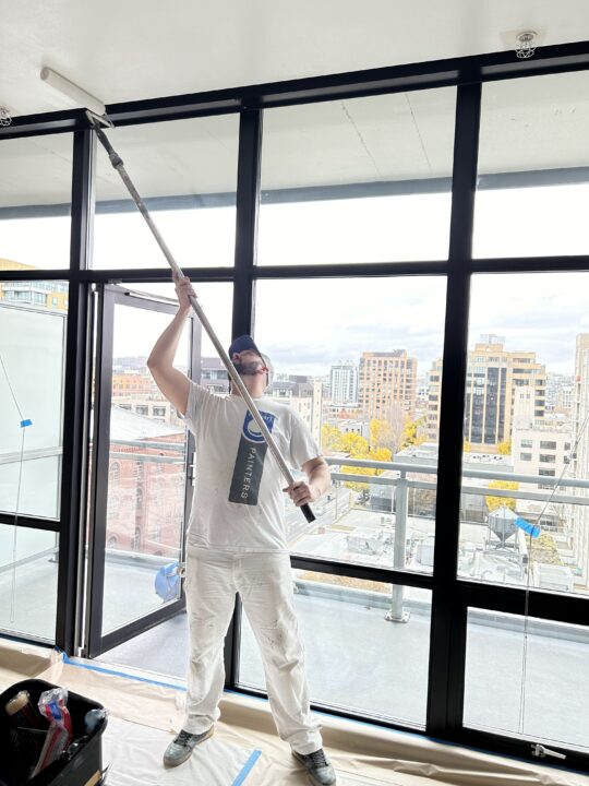 Person in white attire painting a ceiling with a roller on an extended pole in a room with large windows overlooking urban buildings.