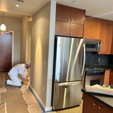 A person in white overalls paints a wall in a kitchen with wooden cabinets and a stainless steel refrigerator. Floor is covered with protective paper.