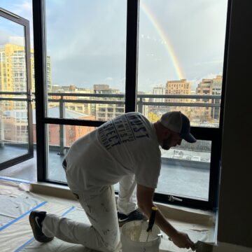 A person in white clothing paints indoors with a brush and bucket, while a rainbow is visible outside through large windows.