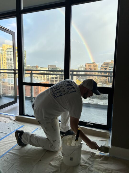 A person in white clothing paints indoors with a brush and bucket, while a rainbow is visible outside through large windows.