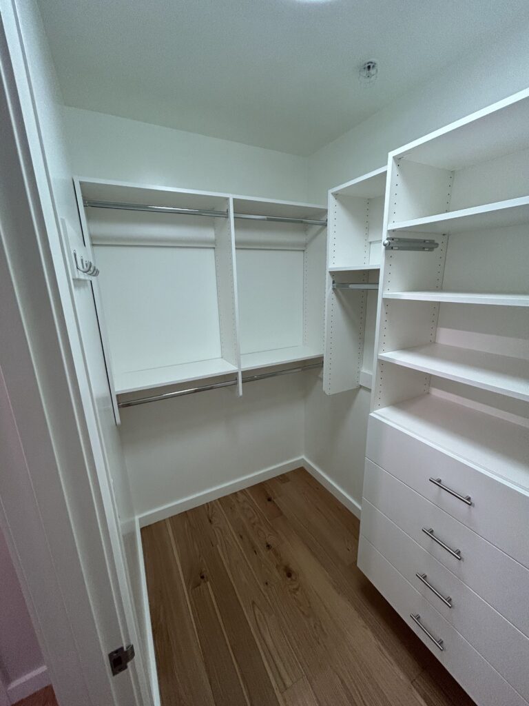 Empty walk-in closet with white shelves, drawers, and hanging rods. The floor is wooden. The walls and ceiling are white.