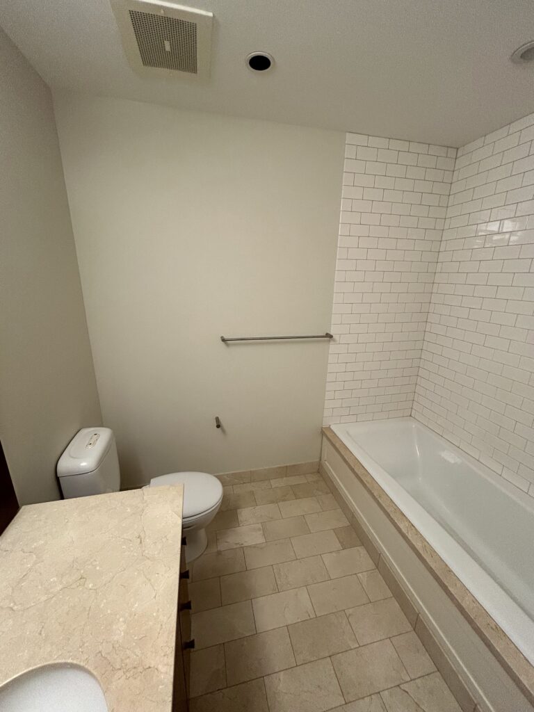 Bathroom with white tile walls, a bathtub, toilet, and marble countertop sink. Beige floor tiles complement the neutral color scheme. A towel bar is mounted above the tub.