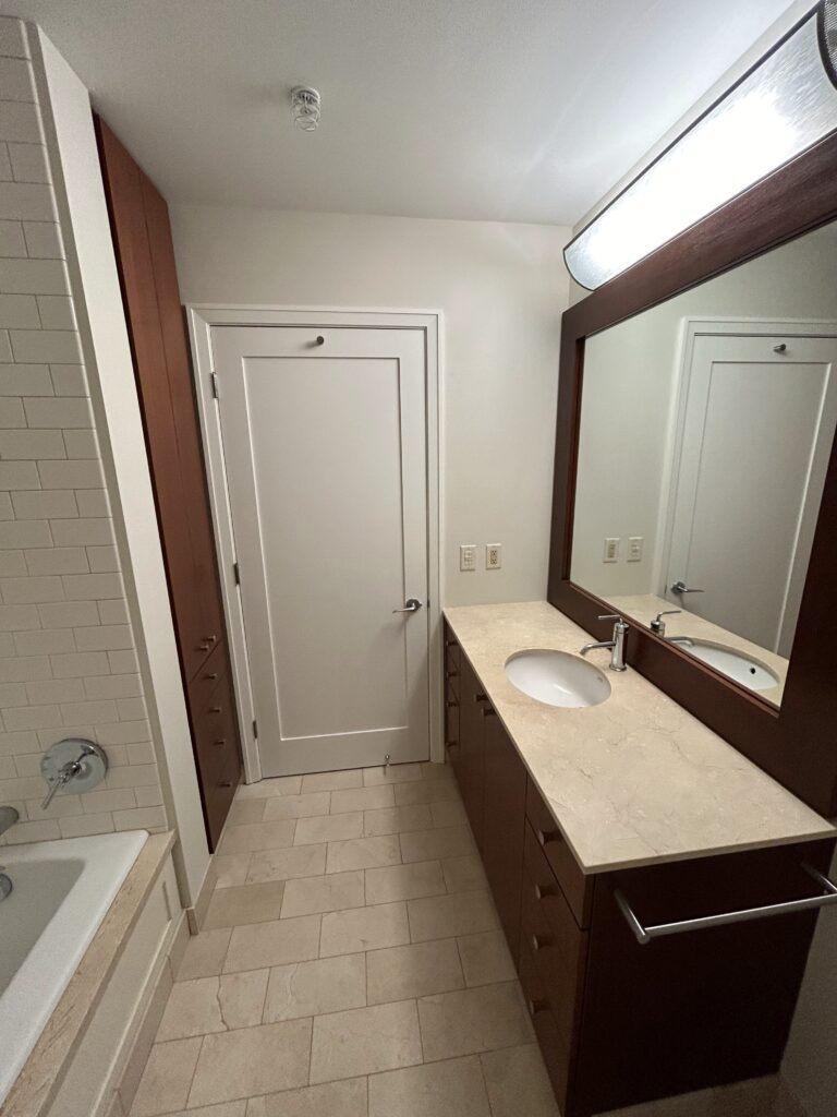 A modern bathroom with a bathtub, a vanity with a sink, and a large mirror. The floor is tiled, and there are wooden cabinets with a door in the background.