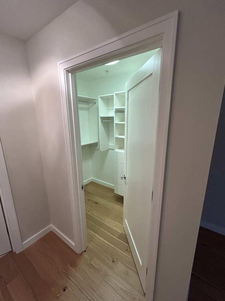 Open door leading to a small, empty walk-in closet with white shelves and wooden flooring.