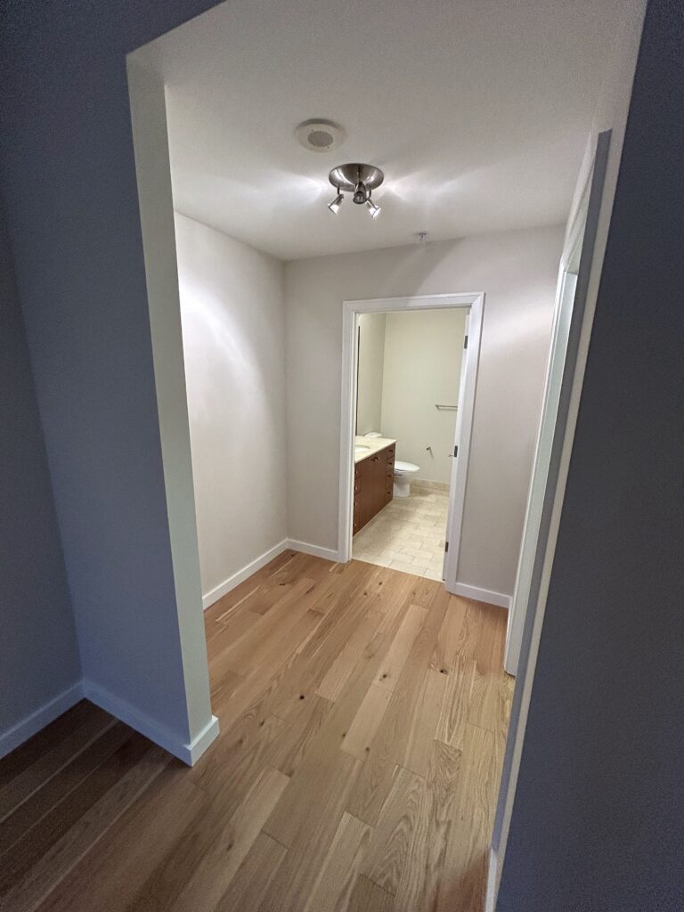 A hallway with a wooden floor leading to a room with a tiled floor. A ceiling light fixture is visible.