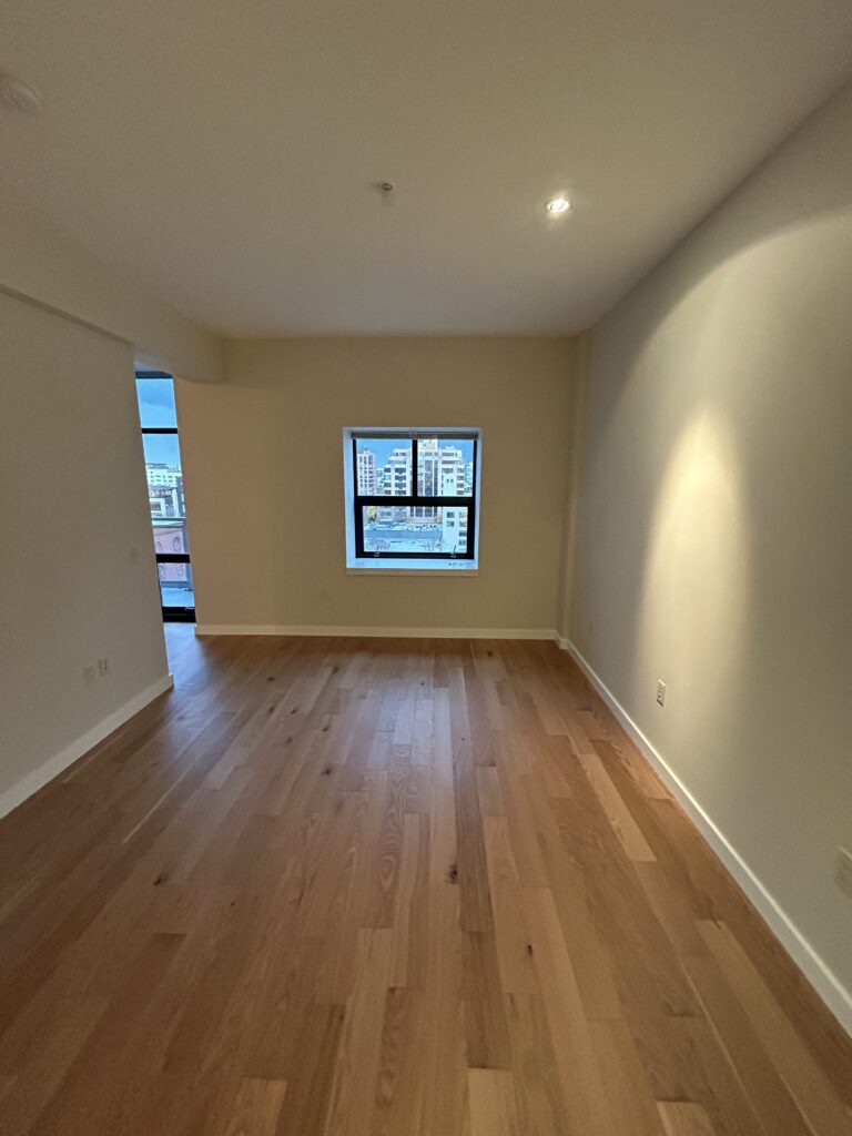 Empty room with light wood flooring and cream walls. A single window shows a cityscape view.