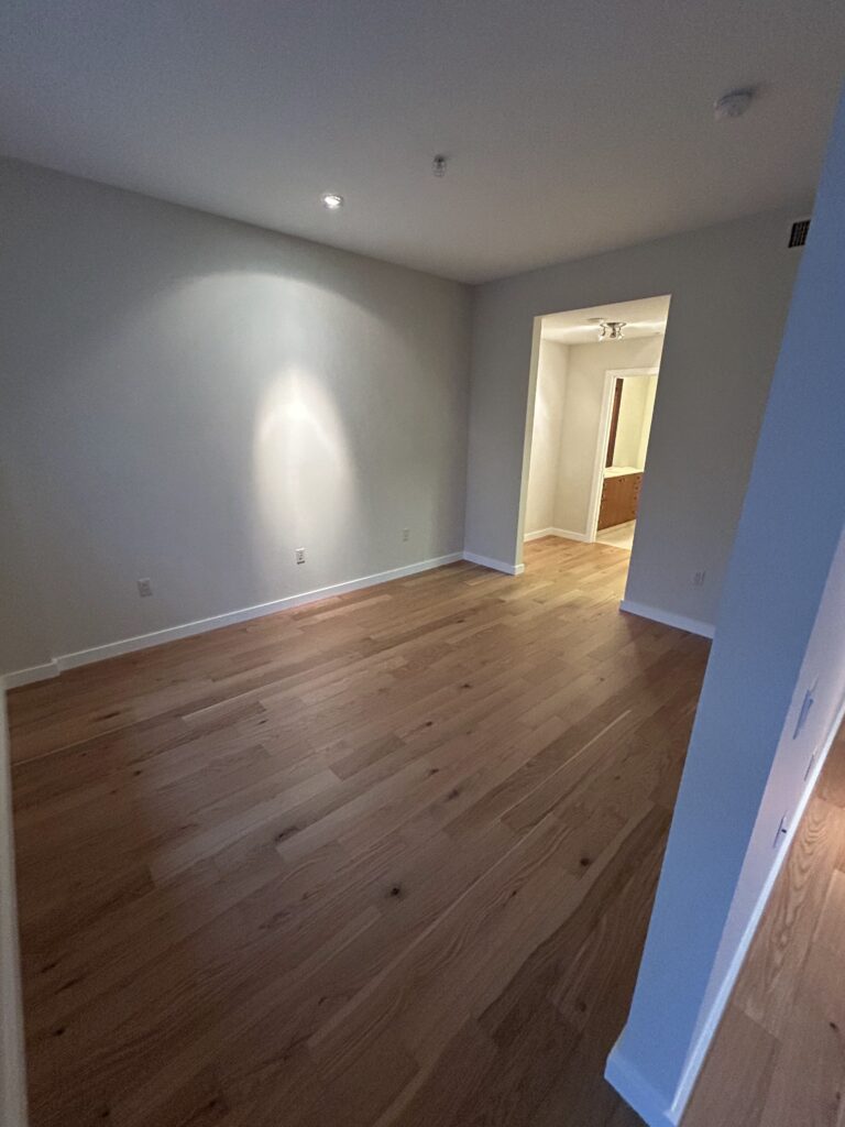 A room with wooden flooring, white walls, and recessed lighting. An open doorway leads to another room with similar flooring and lighting.