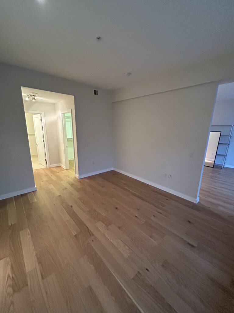 Empty room with light wood flooring, cream walls, and an open doorway leading to a lit hallway and adjacent room.