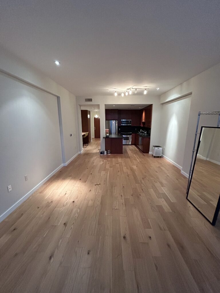 Spacious empty room with light wood flooring, a kitchen area featuring dark cabinetry and appliances at the far end, and a floor mirror on the right.