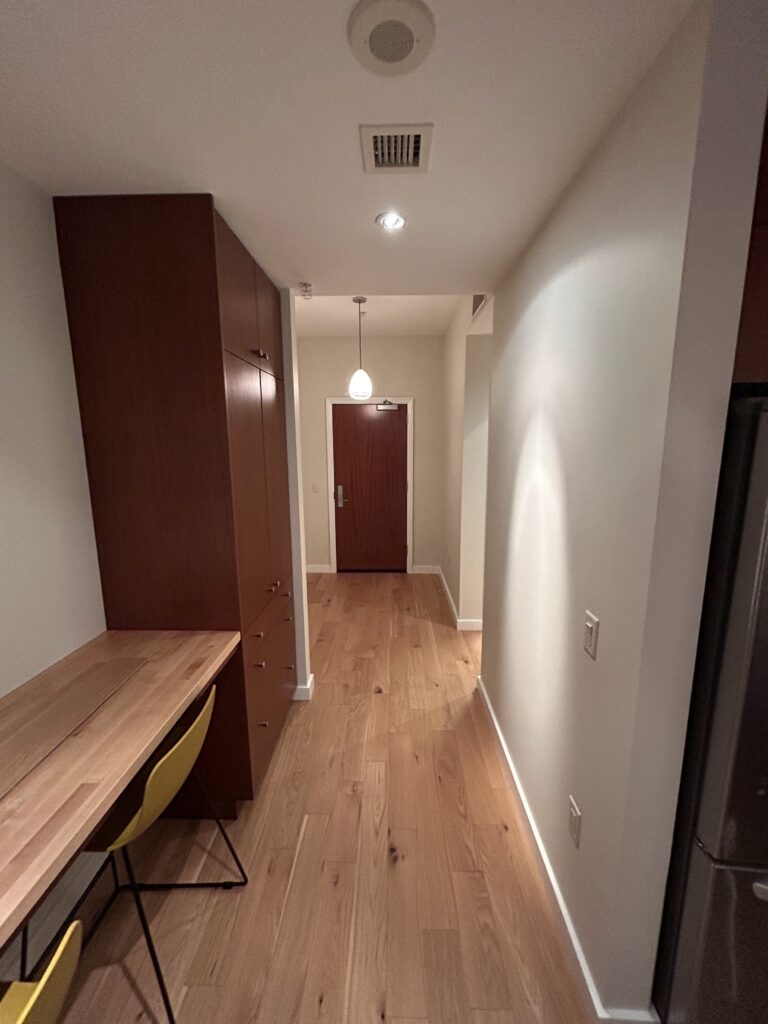 A hallway with wooden flooring features a wooden desk with a yellow chair on the left, a wooden cabinet, and a closed door at the end. A ceiling light fixture hangs in the center.