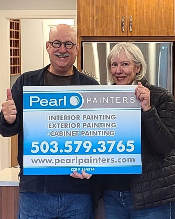 Two older adults indoors hold a Pearl Painters sign for painting services in Portland, showing contact info and website.