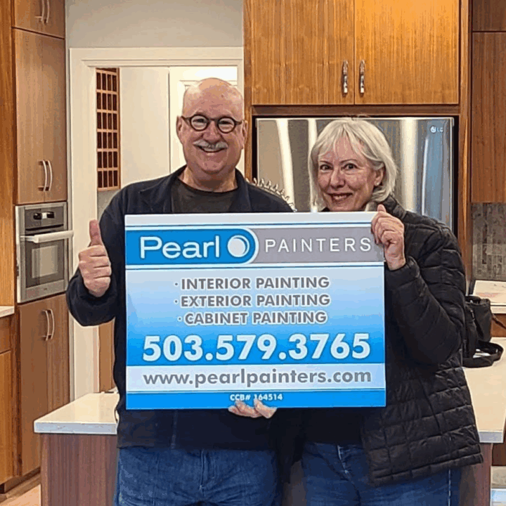 Two people in a kitchen hold a Pearl Painters sign for Portland painter services: interior, exterior, and cabinet painting.