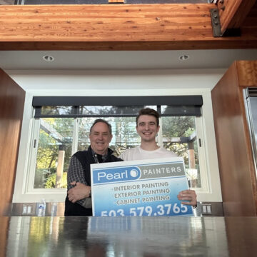 Two painters hold a Pearl Painters sign for painting services indoors. Wood beams and window behind them. Likely in Portland.
